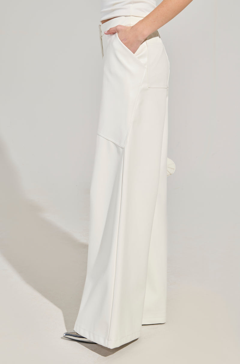 A person wearing the ASHLYN FAUX LEATHER PANT IN WHITE, which features a high-waisted, wide-legged design and smooth texture, stands with their hand in the pocket in a minimalistic setting. The chic silhouette of these pants elegantly complements the partially visible figure from waist down. 3