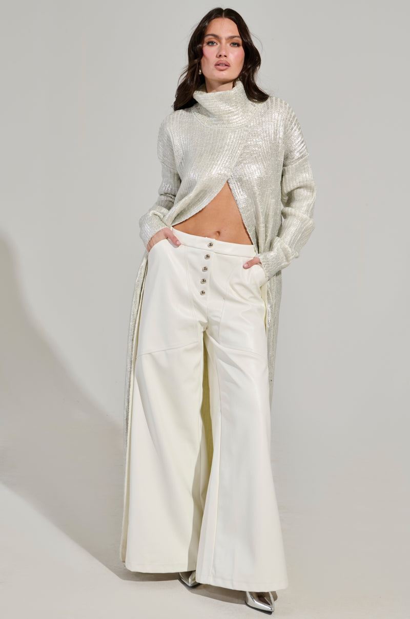 A woman poses in a fashionable ensemble consisting of a long, metallic silver top with a high neckline and front cutout, paired with the ASHLYN FAUX LEATHER PANT IN WHITE. She stands confidently against a neutral background. 6
