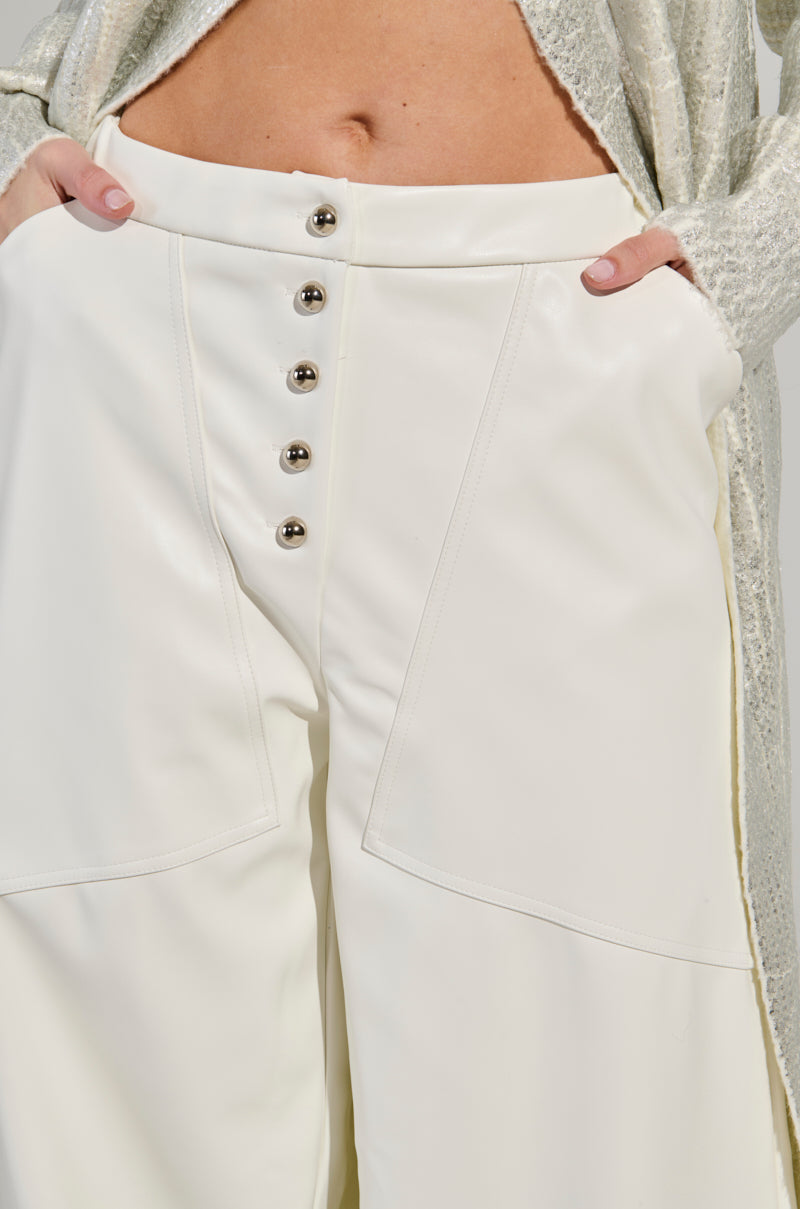 A person wearing the ASHLYN FAUX LEATHER PANT IN WHITE, featuring a mid-rise and wide-leg silhouette with striking metallic buttons down the front. Their hands rest on the waistband of these stylish trousers, seamlessly paired with a light, textured top. 7