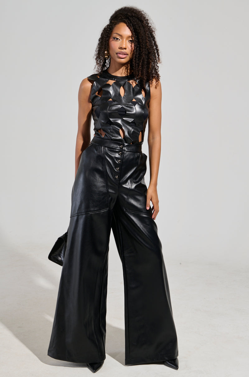 A person with curly hair is wearing a sleek black leather ensemble, featuring a high-rise sleeveless top with cut-out designs. The ASHLYN FAUX LEATHER PANT IN BLACK adds to the look with its wide-leg silhouette, and they hold a small black bag while posing against a plain background.