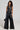 A person with curly hair is wearing a sleek black leather ensemble, featuring a high-rise sleeveless top with cut-out designs. The ASHLYN FAUX LEATHER PANT IN BLACK adds to the look with its wide-leg silhouette, and they hold a small black bag while posing against a plain background.