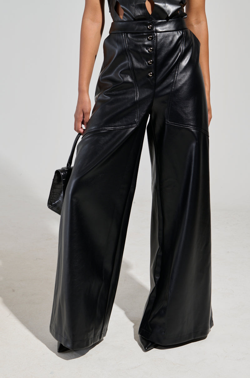 An individual is styled in the ASHLYN FAUX LEATHER PANT IN BLACK, featuring a high-rise, wide-leg design and a button-up front, complemented by a small black handbag. The backdrop is plain and light-colored.
