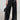 An individual is styled in the ASHLYN FAUX LEATHER PANT IN BLACK, featuring a high-rise, wide-leg design and a button-up front, complemented by a small black handbag. The backdrop is plain and light-colored.