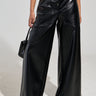 An individual is styled in the ASHLYN FAUX LEATHER PANT IN BLACK, featuring a high-rise, wide-leg design and a button-up front, complemented by a small black handbag. The backdrop is plain and light-colored.