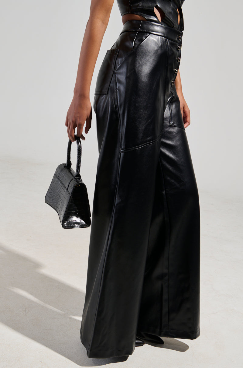 Clad in the Ashlyn Faux Leather Pant in Black, known for its high-rise fit and wide-leg silhouette accented by buttons down the front, the individual is holding a small black handbag with a textured design. The scene is set against a plain and minimalistic background.