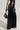 Clad in the Ashlyn Faux Leather Pant in Black, known for its high-rise fit and wide-leg silhouette accented by buttons down the front, the individual is holding a small black handbag with a textured design. The scene is set against a plain and minimalistic background.