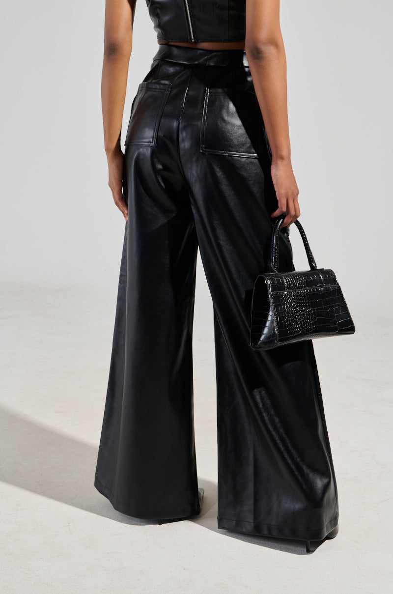 A person wearing the ASHLYN FAUX LEATHER PANT IN BLACK, styled with a matching sleeveless top, holds a small textured black handbag. The ensemble showcases a wide-leg silhouette, viewed from the back against a plain gray background.