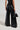A person wearing the ASHLYN FAUX LEATHER PANT IN BLACK, styled with a matching sleeveless top, holds a small textured black handbag. The ensemble showcases a wide-leg silhouette, viewed from the back against a plain gray background.