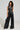 A person wearing a stylish black leather ensemble, highlighting a sleeveless top with cut-out details and the ASHLYN FAUX LEATHER PANT IN BLACK in a wide-leg silhouette. They hold a black handbag and have curly hair, standing against a plain background.