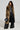 A person with curly hair wears a leopard-print fur coat over a striking black outfit featuring cutouts and the ASHLYN FAUX LEATHER PANT IN BLACK with high-rise, wide-leg design. They hold a black textured handbag and pose confidently against a plain background.