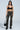 A person poses against a plain backdrop wearing black chunky boots, sunglasses, a choker, arm cuff, and the MAKE A MOVE CAMO PANT featuring a high-rise waist and cargo pockets. They pair it with a black crop top and hold a black handbag at their side.