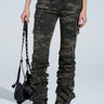 A person wearing MAKE A MOVE CAMO PANT with ruched legs and black chunky boots holds a small black bag against a plain, light-colored background.