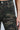 A close-up of a person wearing MAKE A MOVE CAMO PANT, high-rise with camo pattern and cargo-style pockets. Their left hand rests on the side, revealing part of a forearm tattoo. The pants feature silver button details.