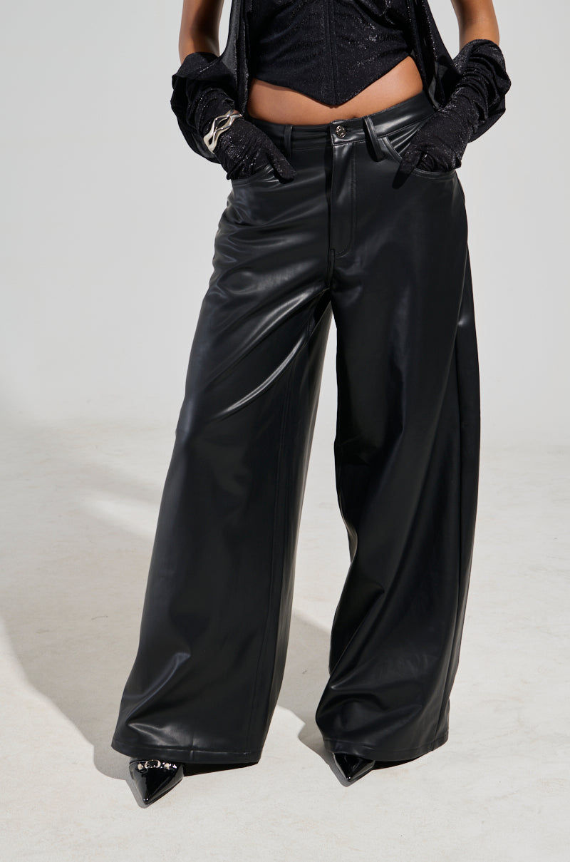 A person is showcasing the GABRIELLE Wide Leg Faux Leather Pants in black paired with a black corset-style top. Their hands, clad in sleek black gloves, rest in the pants' pockets. The minimal background enhances this edgy fashion statement.