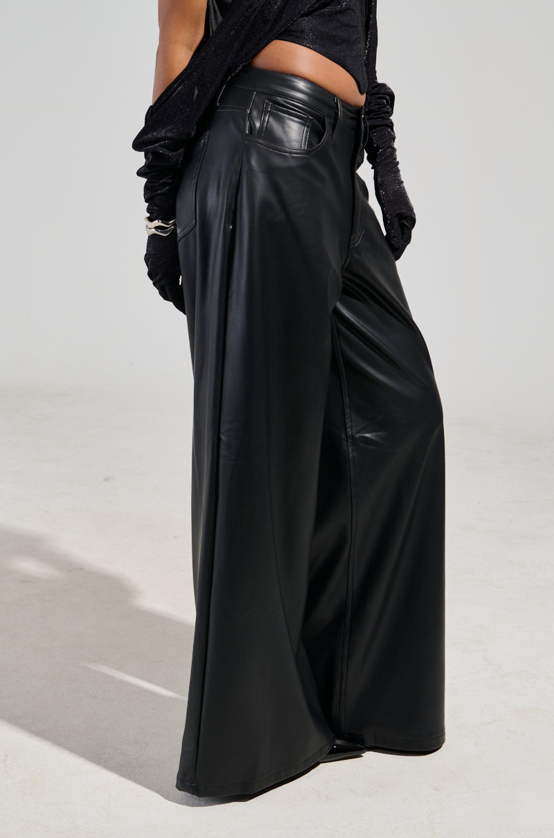 A person wearing the GABRIELLE wide-leg faux leather pants in black and a matching top, with gloved hands visible against a plain, light-colored background.