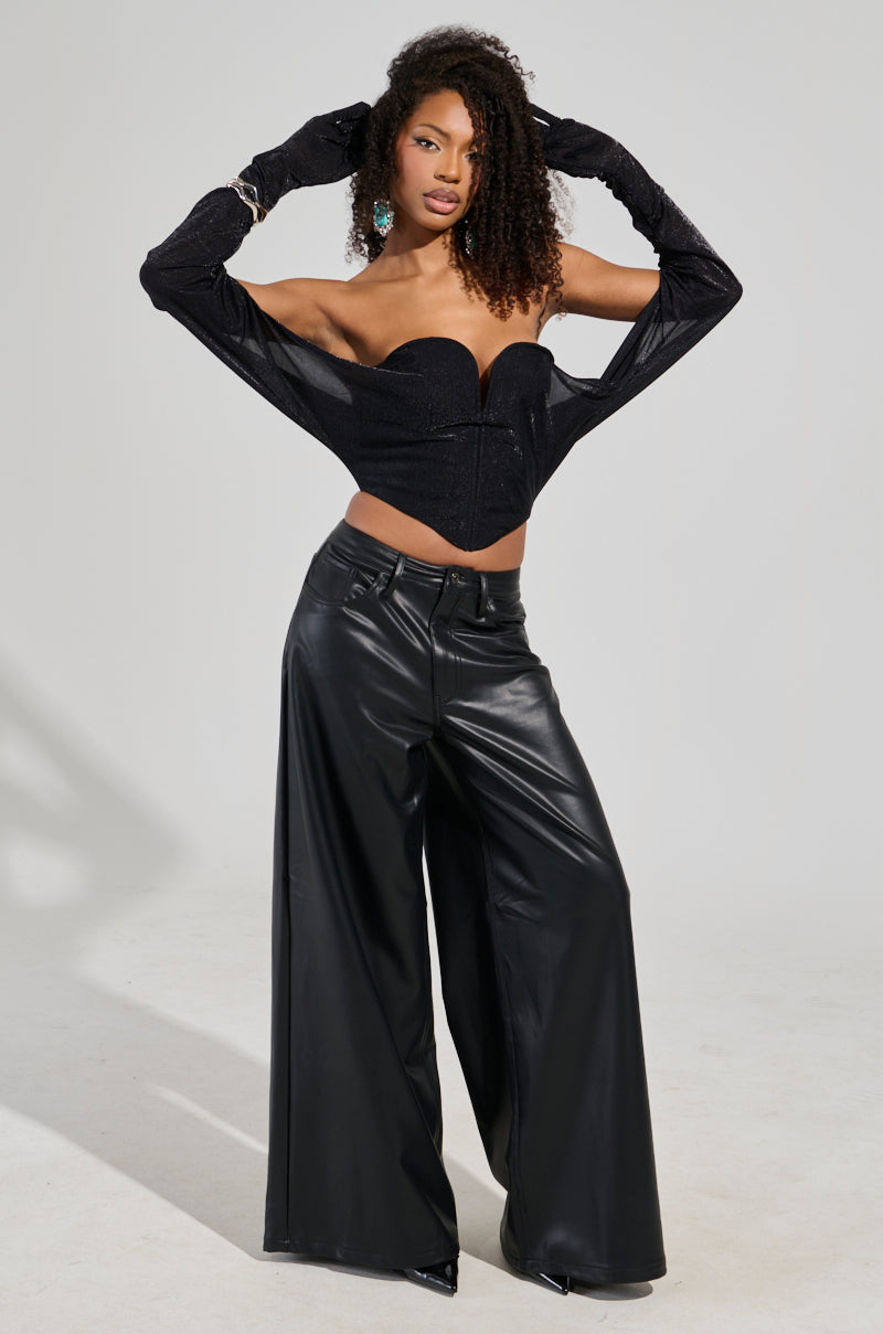 A person confidently poses wearing a black, strapless corset top paired with the Gabrielle Wide Leg Faux Leather Pant in black. They have long curly hair and are accessorized with gloves and earrings, standing against a plain background.