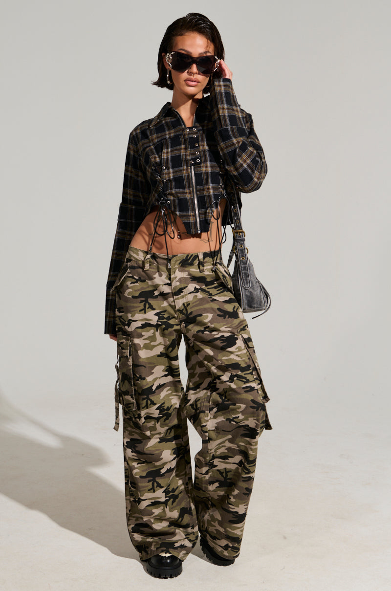 A person wearing sunglasses poses confidently in a plaid crop top and STREET STYLE WIDE LEG CAMO PANT, holding a purse. The background is a plain, light color, showcasing their street style fashion sense.