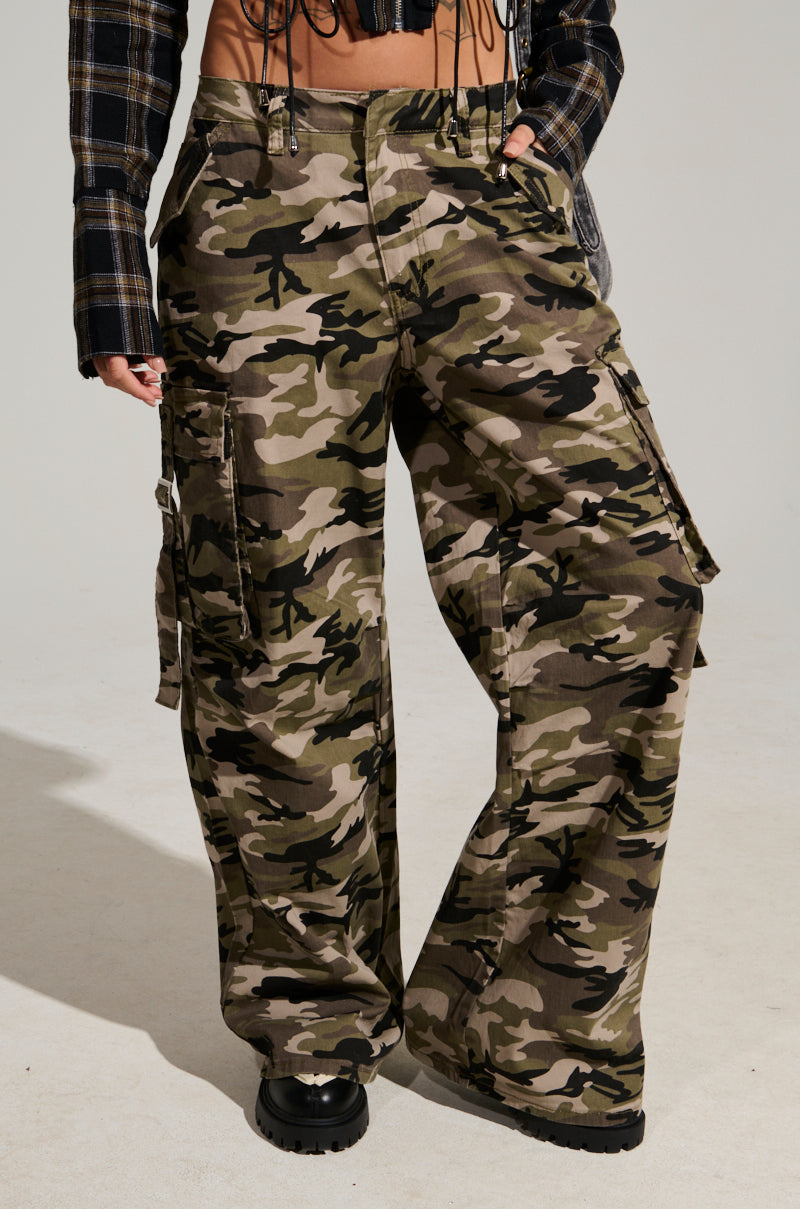Wearing STREET STYLE WIDE LEG CAMO PANT and black boots, a person confidently showcases street style in a relaxed pose. The pants feature large pockets, and the addition of a plaid jacket enhances the look. A plain, light-colored background perfectly complements the ensemble.