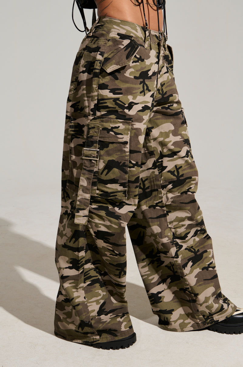 A person is rocking the street style with the STREET STYLE WIDE LEG CAMO PANT featuring large pockets and black sneakers. The green, brown, and beige camouflage pattern adds a bold flair, set against a plain, light-colored background.