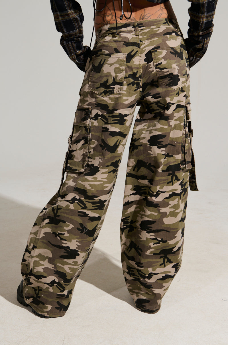 A person displays street style in the STREET STYLE WIDE LEG CAMO PANT, featuring multiple pockets, viewed from behind. They're standing on a light-colored surface against a neutral background, with hands confidently resting on their hips.