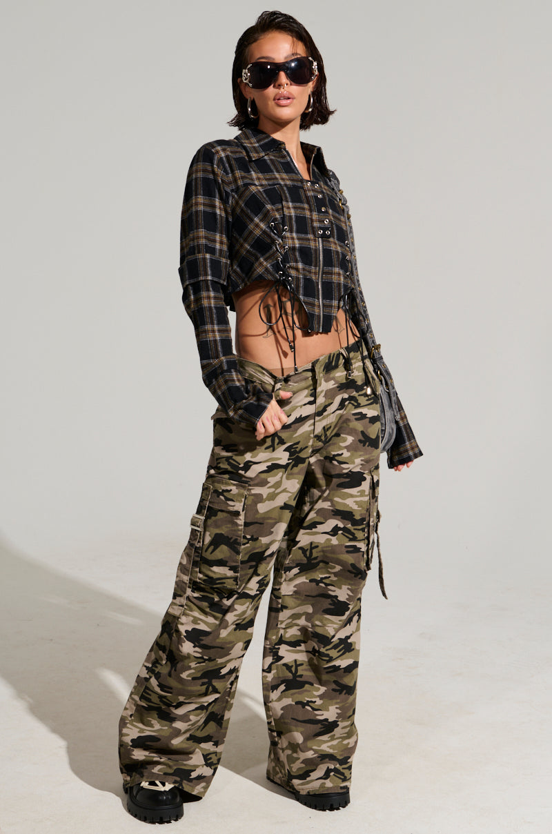 A person showcases their street style by posing in STREET STYLE WIDE LEG CAMO PANTS, complemented by sunglasses, a plaid crop top with lace-up details, and accessorized with short hair and a sleek black crossbody bag against a plain background.