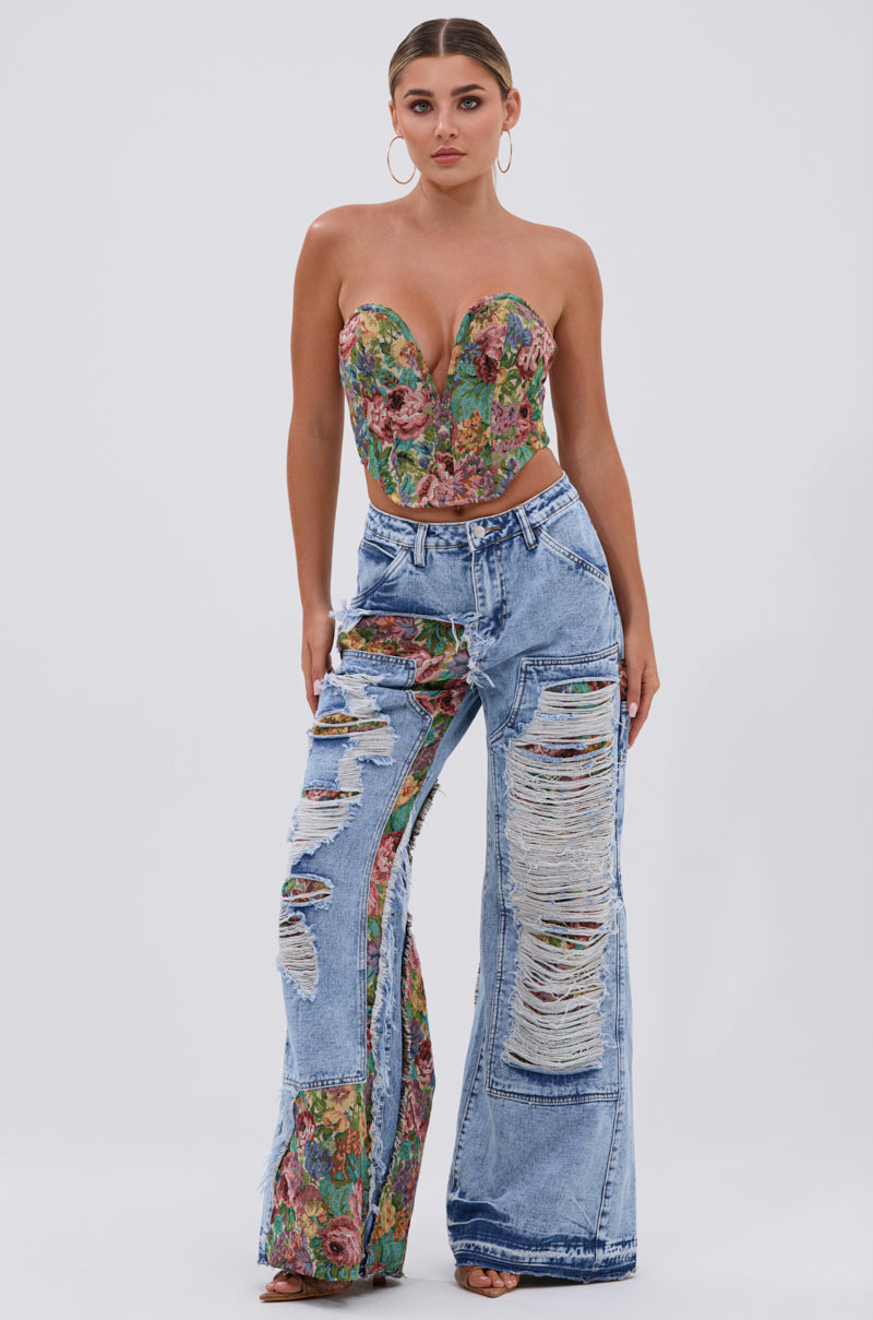 A woman poses confidently against a plain backdrop in the THOUGHT I KNEW YOU RELAXED DENIM WITH BROCADE—high-waisted, wide-leg jeans featuring large rips and floral jacquard panels—paired with a strapless floral bustier and hoop earrings.