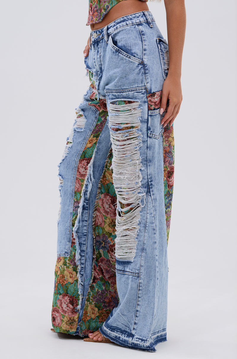 A person stands barefoot against a plain white background, wearing the THOUGHT I KNEW YOU RELAXED DENIM WITH BROCADE—high-waisted wide-leg jeans featuring large distressed rips and floral brocade fabric panels.