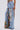 A person stands barefoot against a plain white background, wearing the THOUGHT I KNEW YOU RELAXED DENIM WITH BROCADE—high-waisted wide-leg jeans featuring large distressed rips and floral brocade fabric panels.