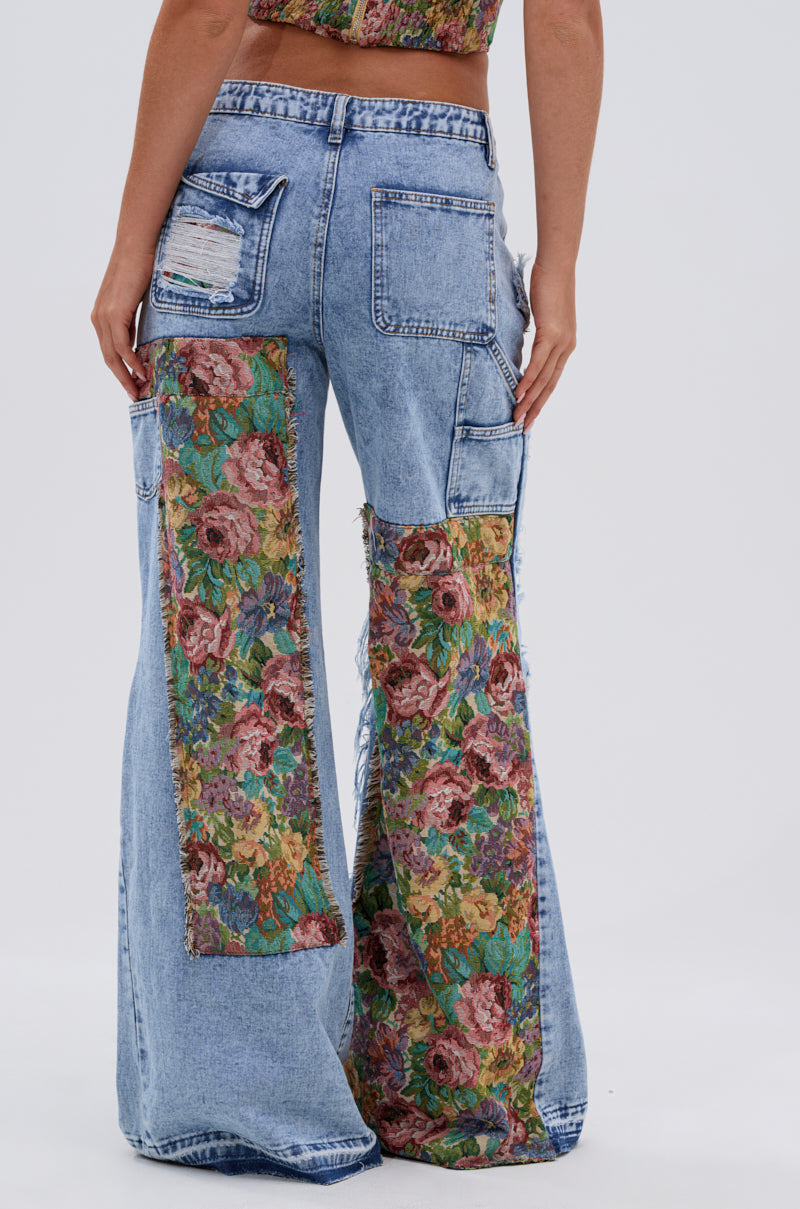 Someone wears the THOUGHT I KNEW YOU RELAXED DENIM WITH BROCADE—high-waisted, wide-leg light blue jeans with large floral brocade patches and multiple pockets—paired with a matching floral crop top. The photo is taken from behind.