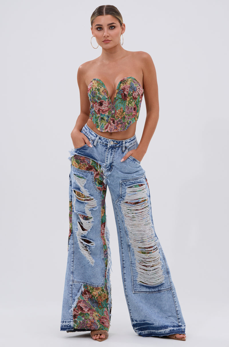 A woman models the "THOUGHT I KNEW YOU RELAXED DENIM WITH BROCADE"—high-waisted, wide-leg distressed jeans featuring floral brocade patches peeking through rips. She stands with hands in pockets against a plain background.