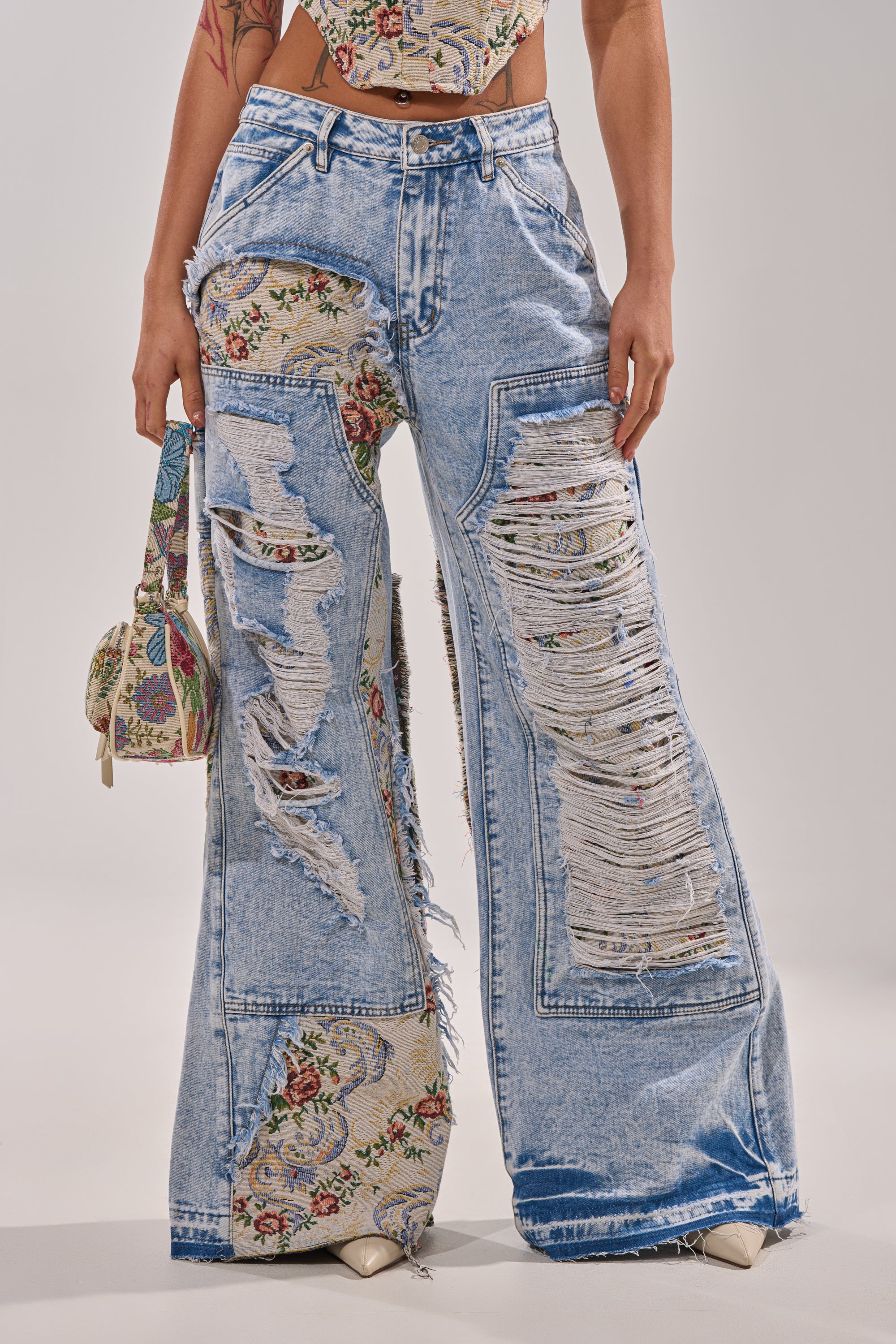 A person wears LANA DISTRESSED JEANS WITH JACQUARD DETAIL, featuring a super-wide silhouette and large ripped patches that reveal floral fabric, styled with a matching floral handbag and sleeveless top against a light plain background. 1