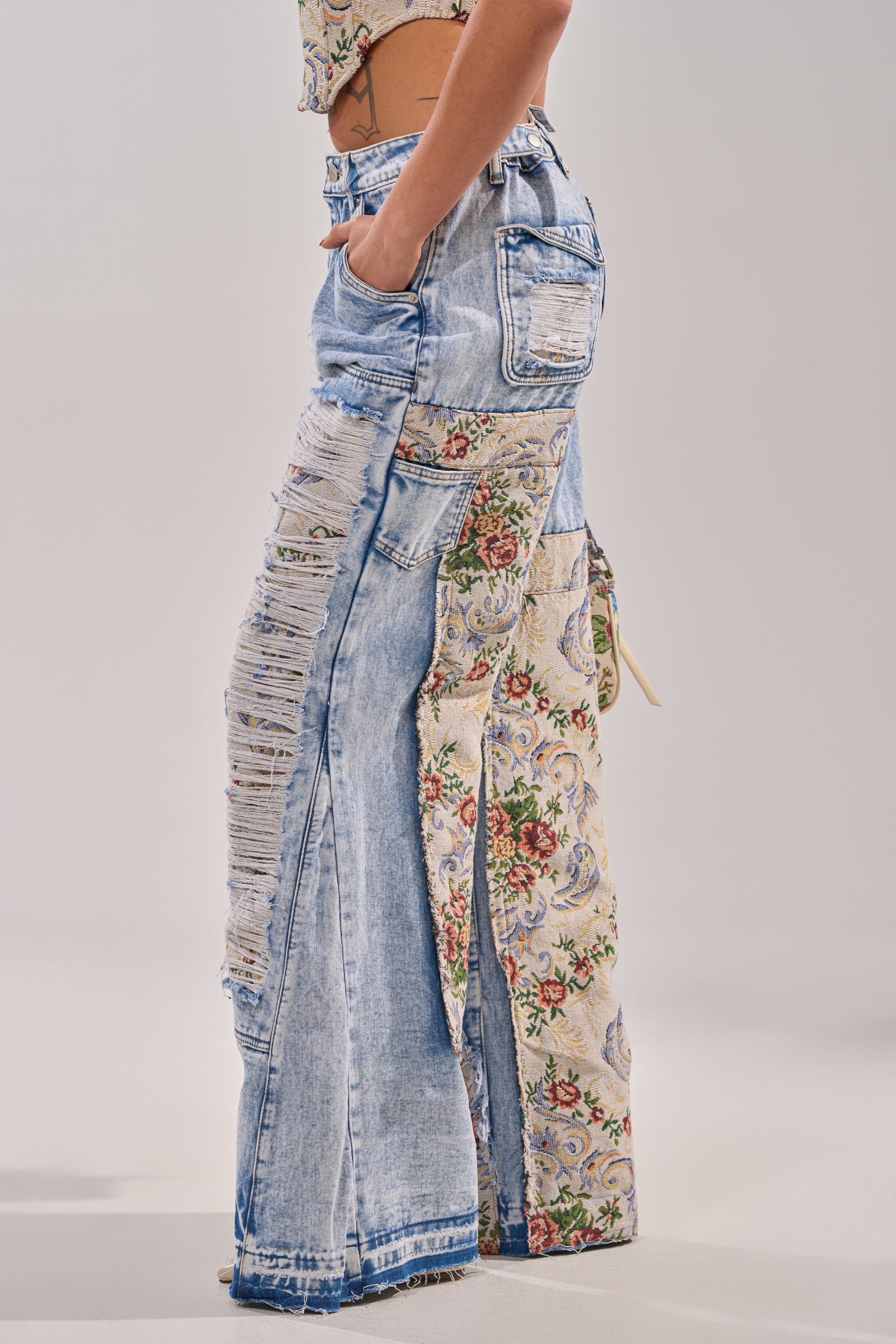 A person wears LANA DISTRESSED JEANS WITH JACQUARD DETAIL—super-wide jeans featuring distressed denim and floral jacquard panels—standing with one hand in their pocket against a plain background. 3