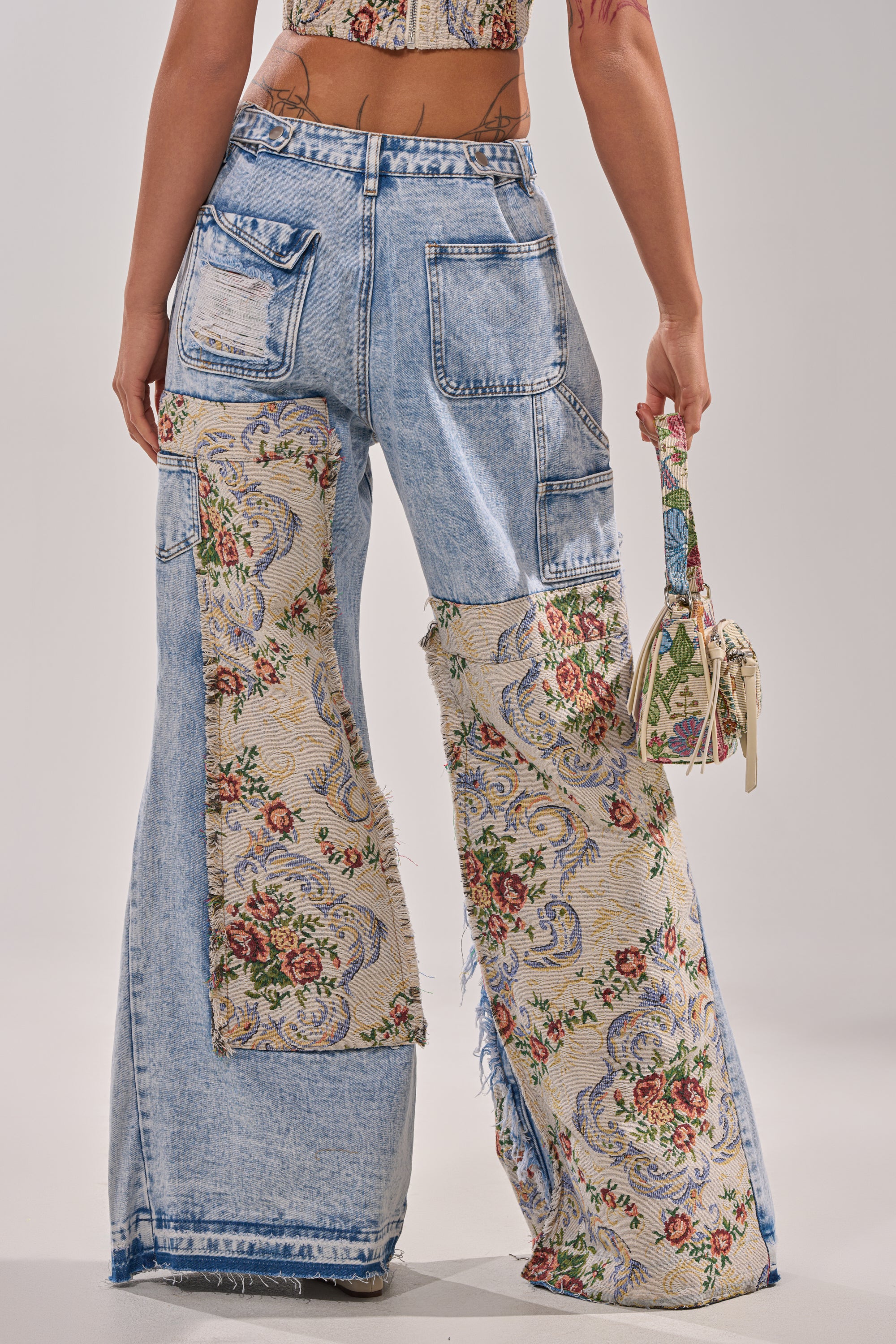 A person faces away, wearing LANA DISTRESSED JEANS WITH JACQUARD DETAIL—light blue denim with floral tapestry patchwork, frayed hems, and a ripped pocket—while holding a small floral-pattern purse. 4