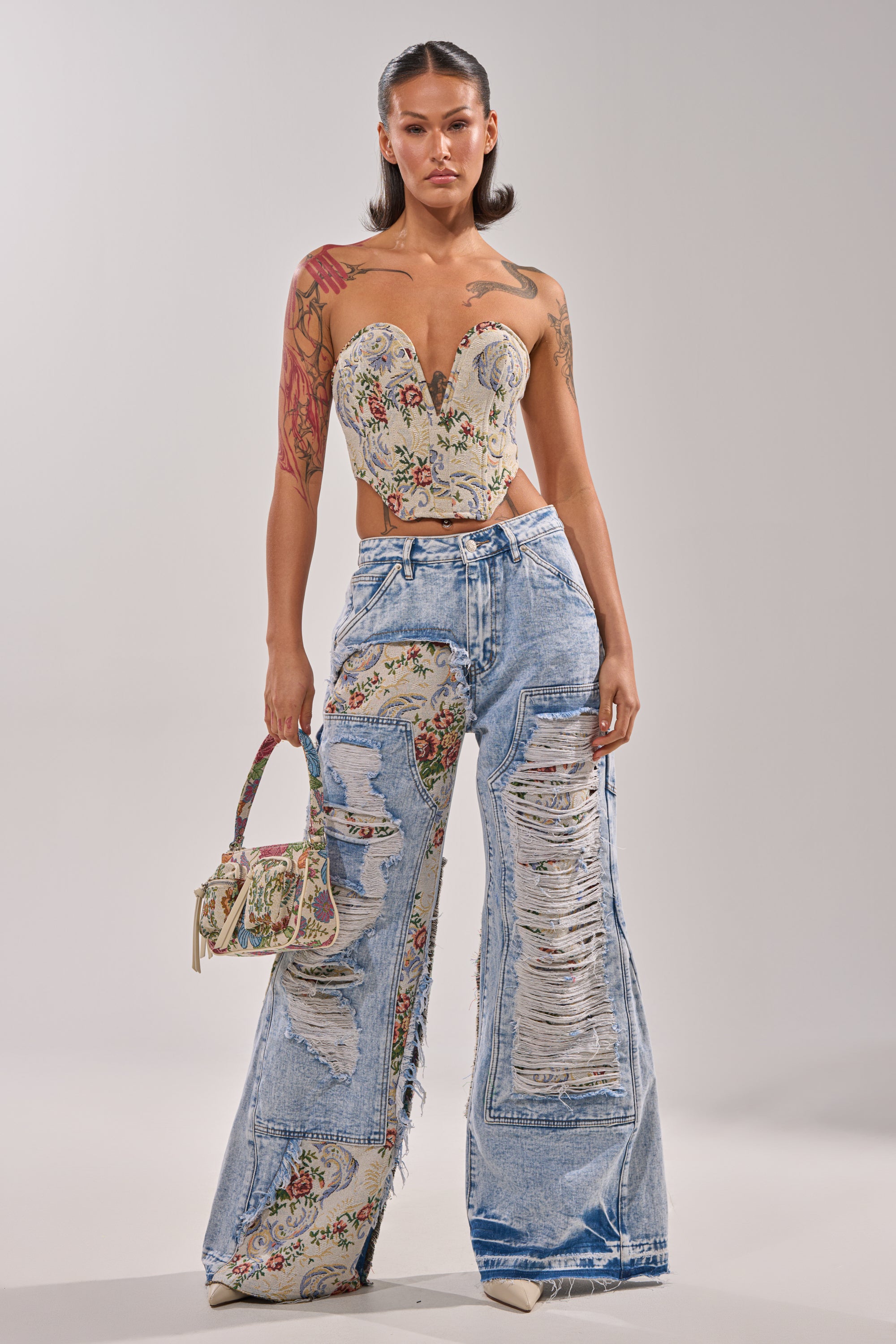 A woman stands confidently in a floral corset top, LANA DISTRESSED JEANS WITH JACQUARD DETAIL, and beige heels, holding a small matching floral handbag. Her hair is sleek and styled back. 7