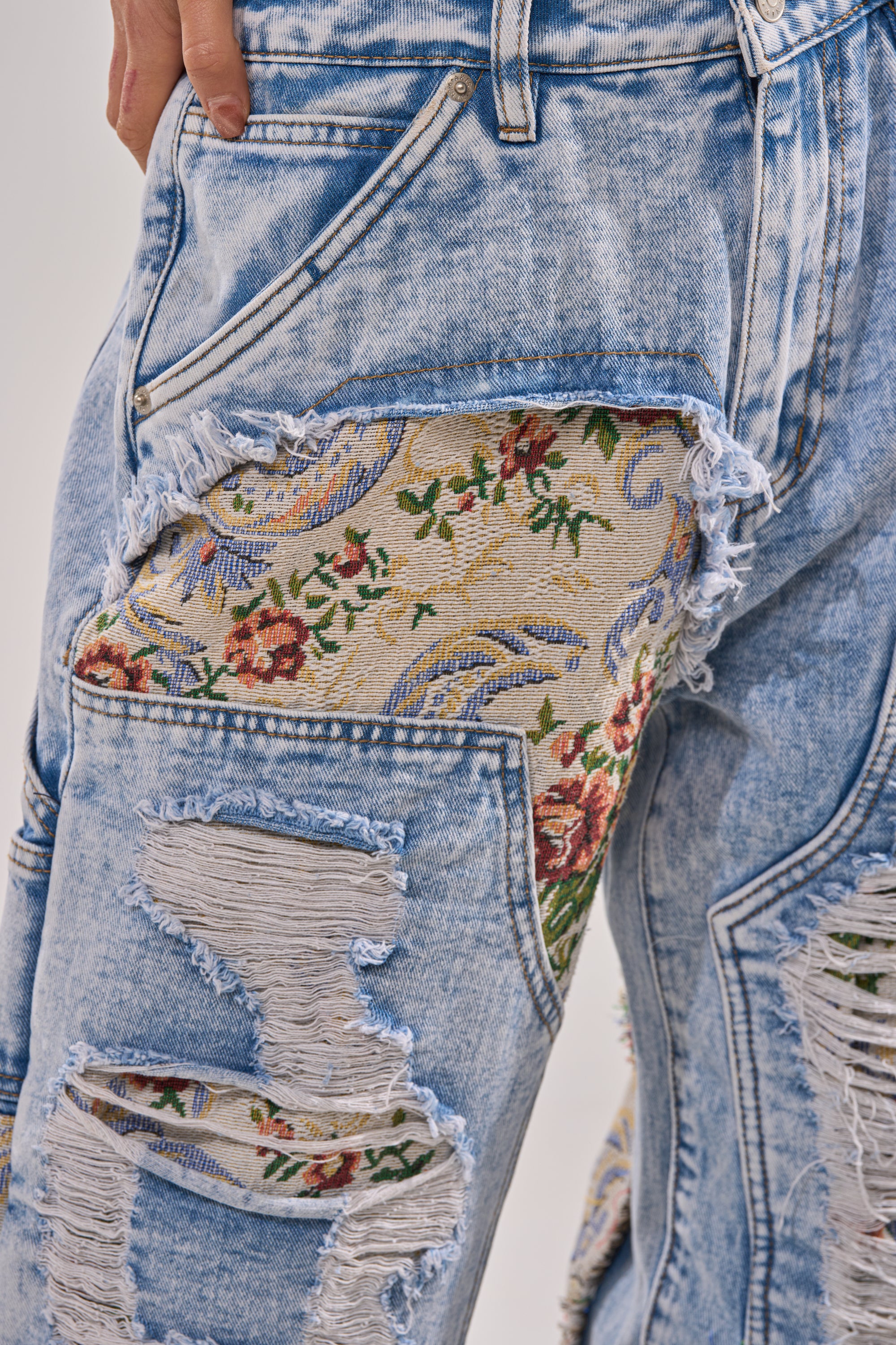 Close-up of the LANA DISTRESSED JEANS WITH JACQUARD DETAIL featuring wide legs, large rips that reveal floral tapestry jacquard fabric beneath, and a hand resting in the front pocket. 8
