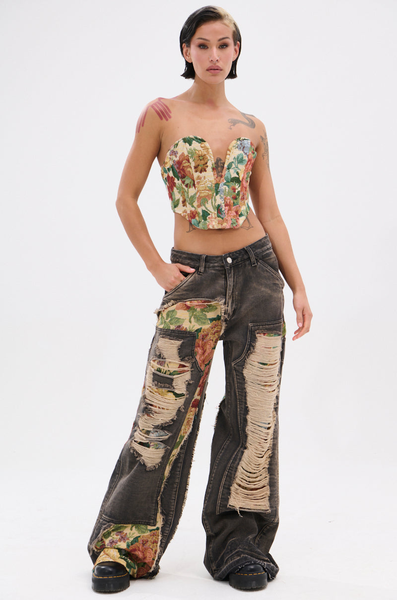 A woman with short dark hair stands confidently in BRIGETTE DISTRESSED JEANS WITH JACQUARD DETAIL, a floral corset top, and black shoes. The jeans' jacquard design peeks through the rips as she poses against a plain white background.