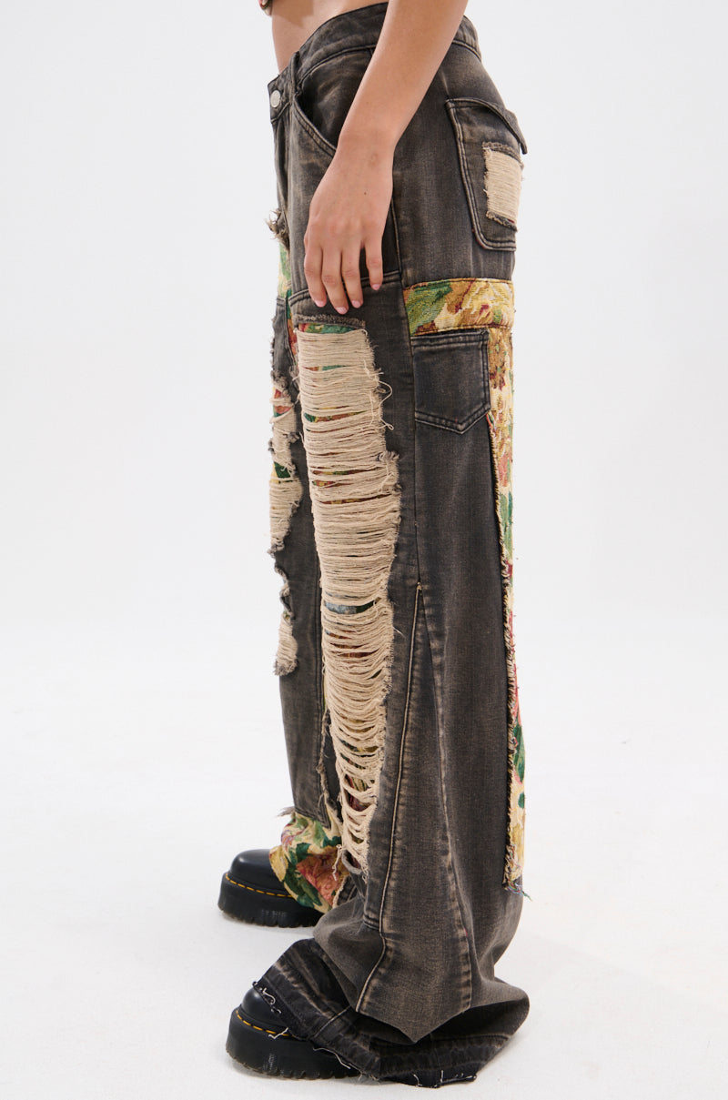 A person stands sideways in BRIGETTE DISTRESSED JEANS WITH JACQUARD DETAIL—high-waisted, dark-wash jeans with large rips and jacquard patches. Black shoes finish the look against a light background.