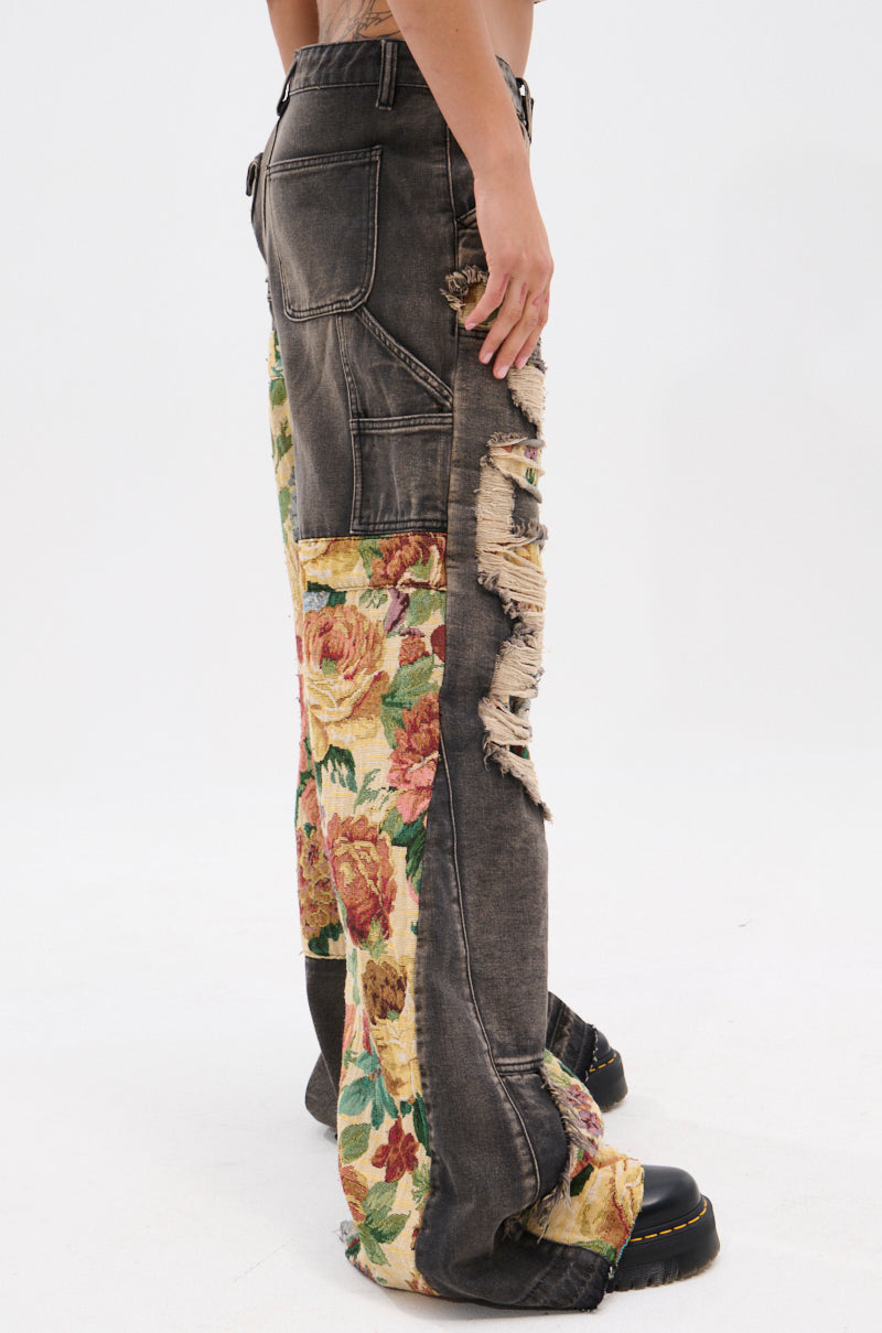 A person wears the BRIGETTE DISTRESSED JEANS WITH JACQUARD DETAIL—patchwork pants featuring distressed denim and floral tapestry accents—paired with black boots, standing against a plain white background.