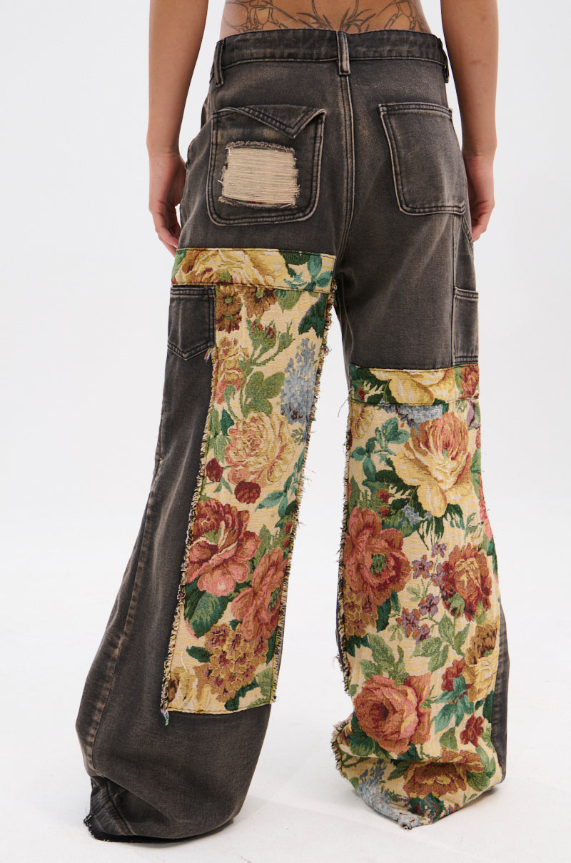 A person wears BRIGETTE DISTRESSED JEANS WITH JACQUARD DETAIL—high-waisted, wide-leg dark denim with colorful floral jacquard and distressed patches, including a frayed back pocket. Their lower back tattoos are visible.