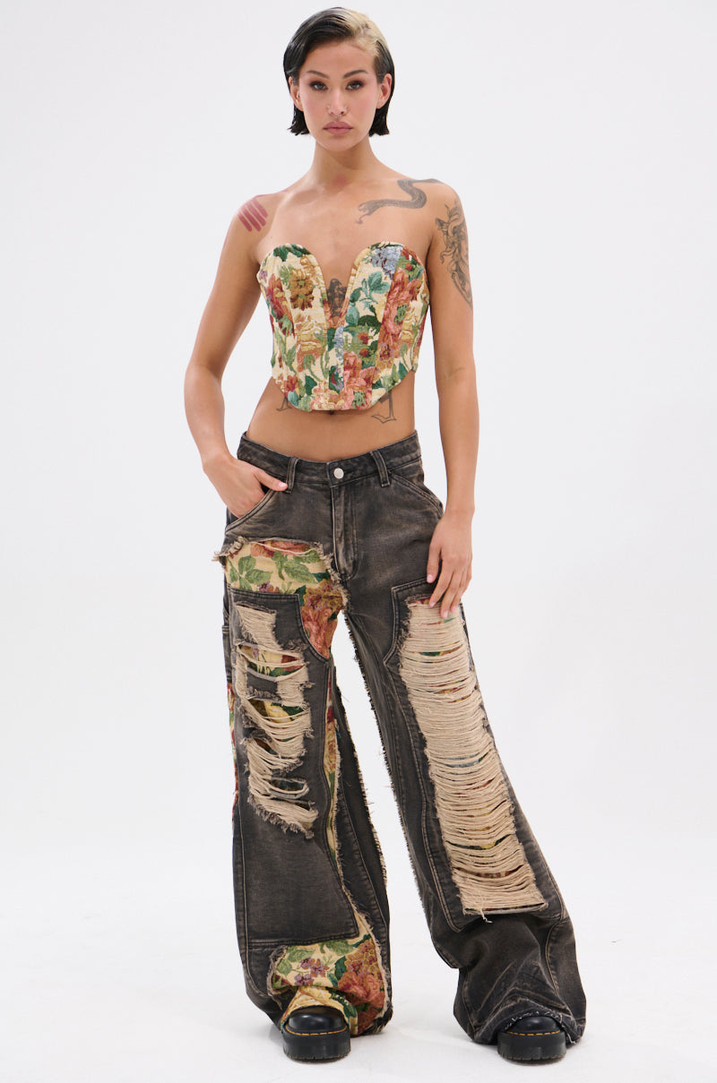 A person stands confidently in BRIGETTE DISTRESSED JEANS WITH JACQUARD DETAIL, featuring wide legs and floral jacquard patches, paired with a floral corset top against a plain white background.