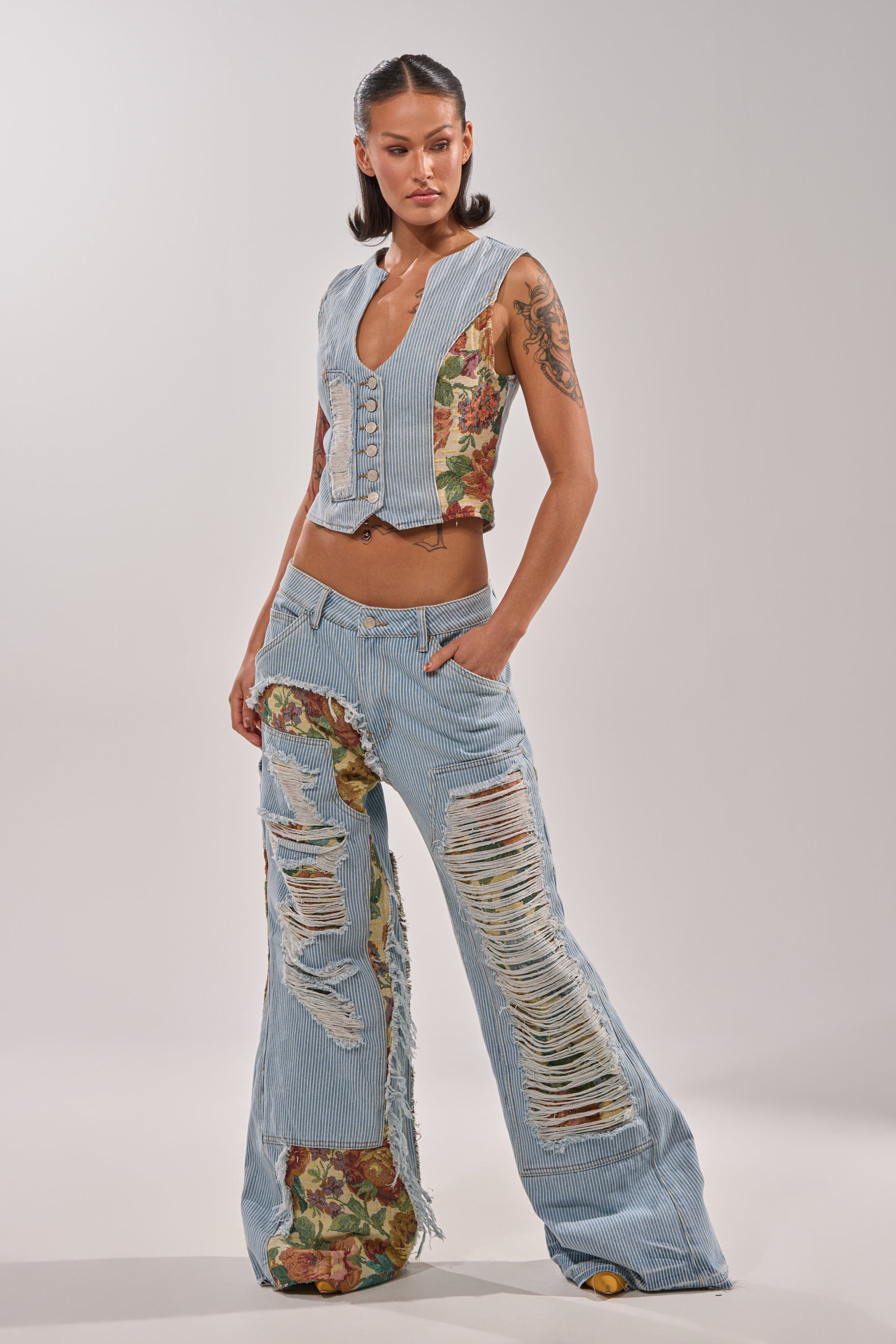 A woman models the GUESS WHO STRIPE DENIM WITH BROCADE—ultra-wide leg, pinstriped jeans with large torn sections and floral brocade patches—paired with a sleeveless vest, against a plain light background. 0