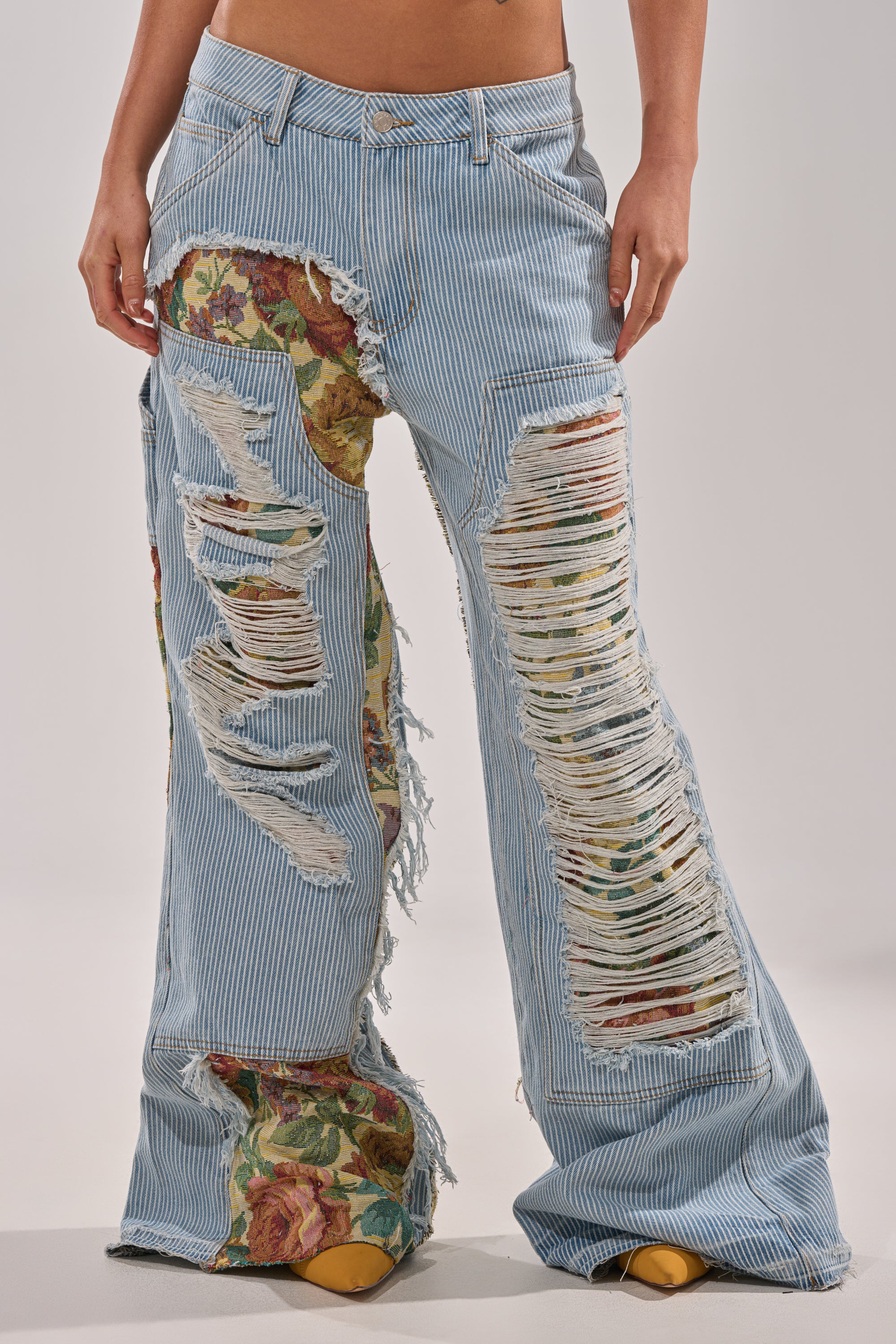 A person wears GUESS WHO STRIPE DENIM WITH BROCADE—light blue, distressed jeans with large rips revealing floral fabric—paired with yellow pointed-toe shoes. Only the lower torso and legs are visible in the image. 1