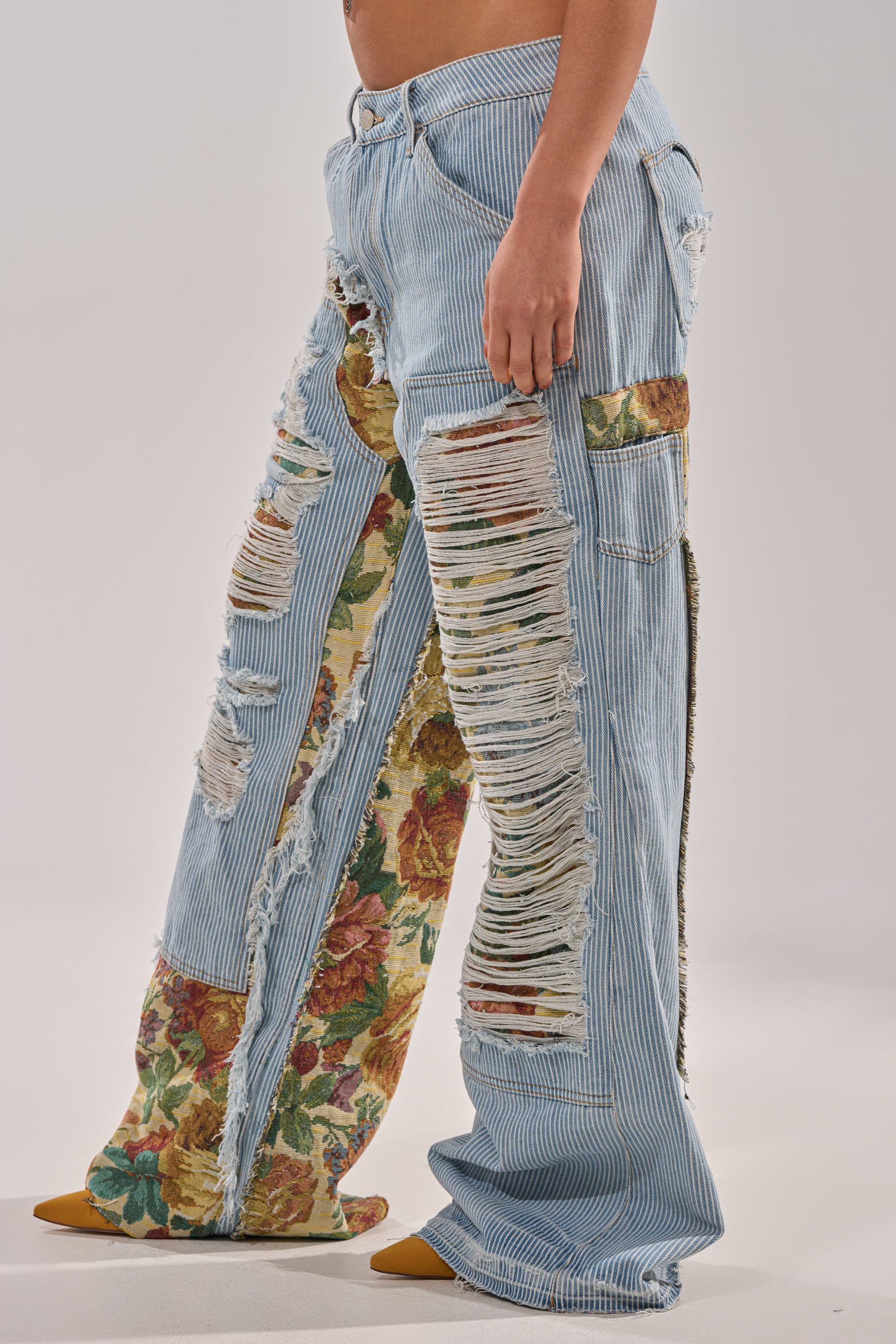 A person wears the GUESS WHO STRIPE DENIM WITH BROCADE, featuring light blue, distressed wide leg jeans with vertical rips and floral brocade patches, standing barefoot against a plain background. 2