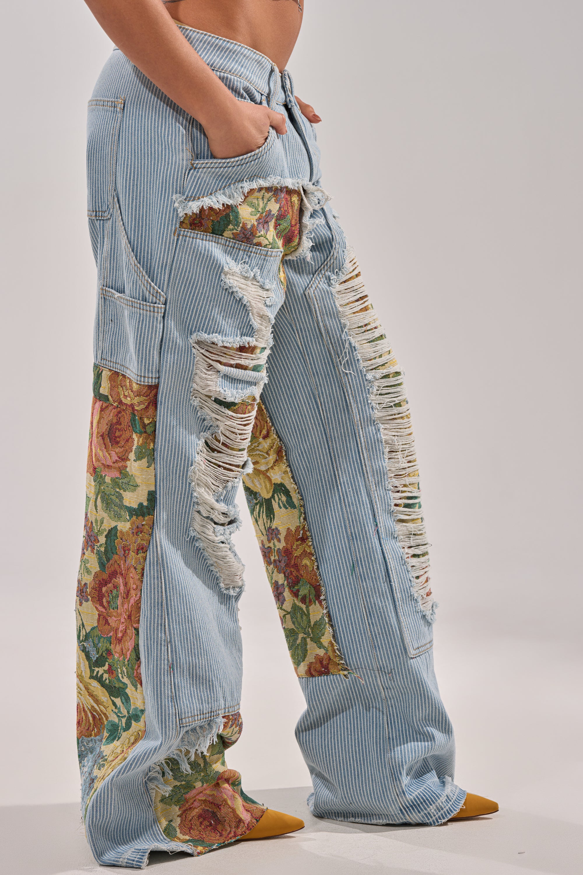 Wearing the GUESS WHO STRIPE DENIM WITH BROCADE, a model stands sideways with one hand in their pocket. The ultra-wide leg jeans feature blue and white stripes, bold distressed details, and floral brocade fabric patches. 3