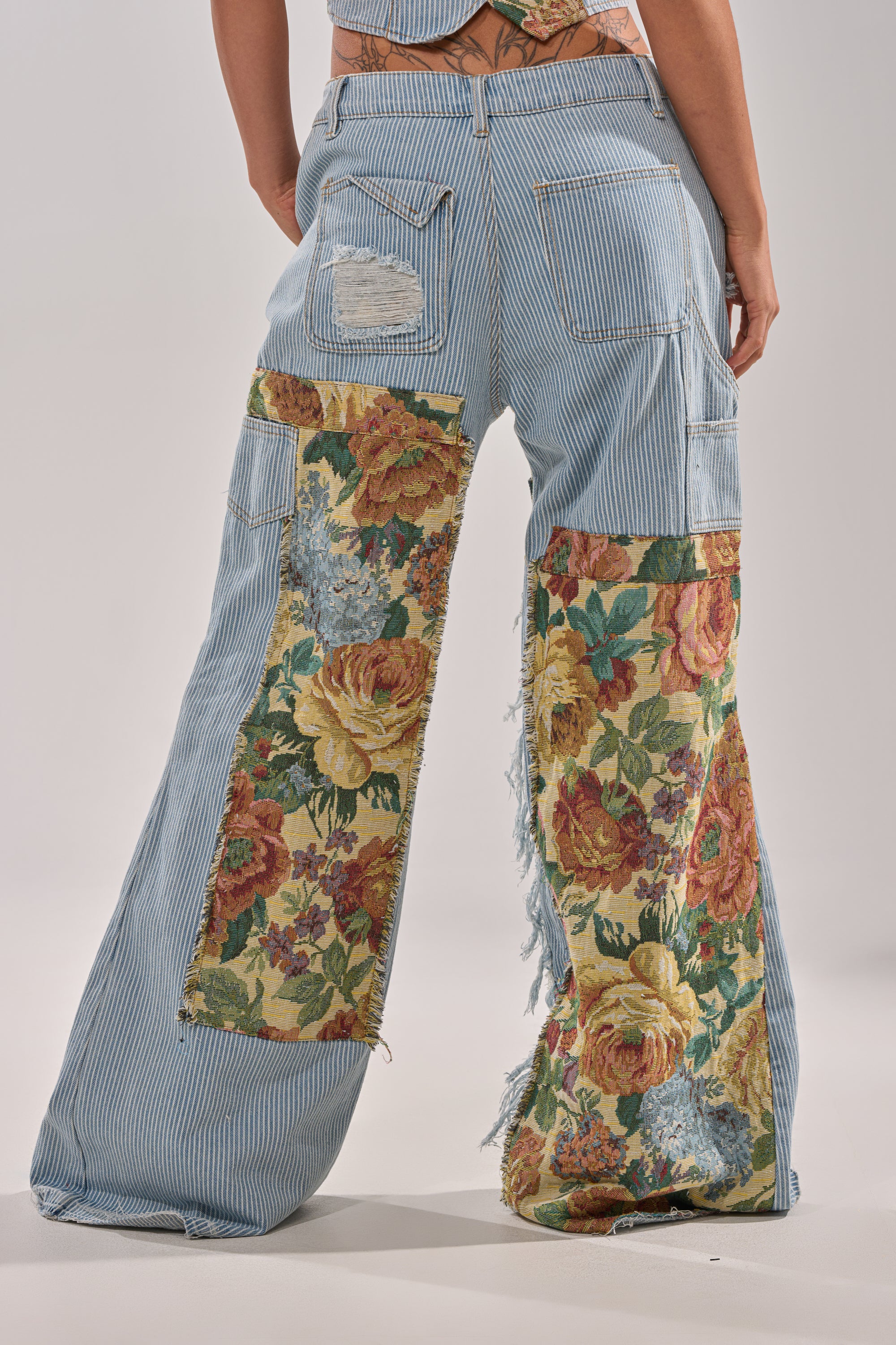 Someone shown from the waist down and viewed from behind is wearing GUESS WHO STRIPE DENIM WITH BROCADE—ultra-wide pinstriped jeans with large floral brocade patches on the back and distressed details. 4