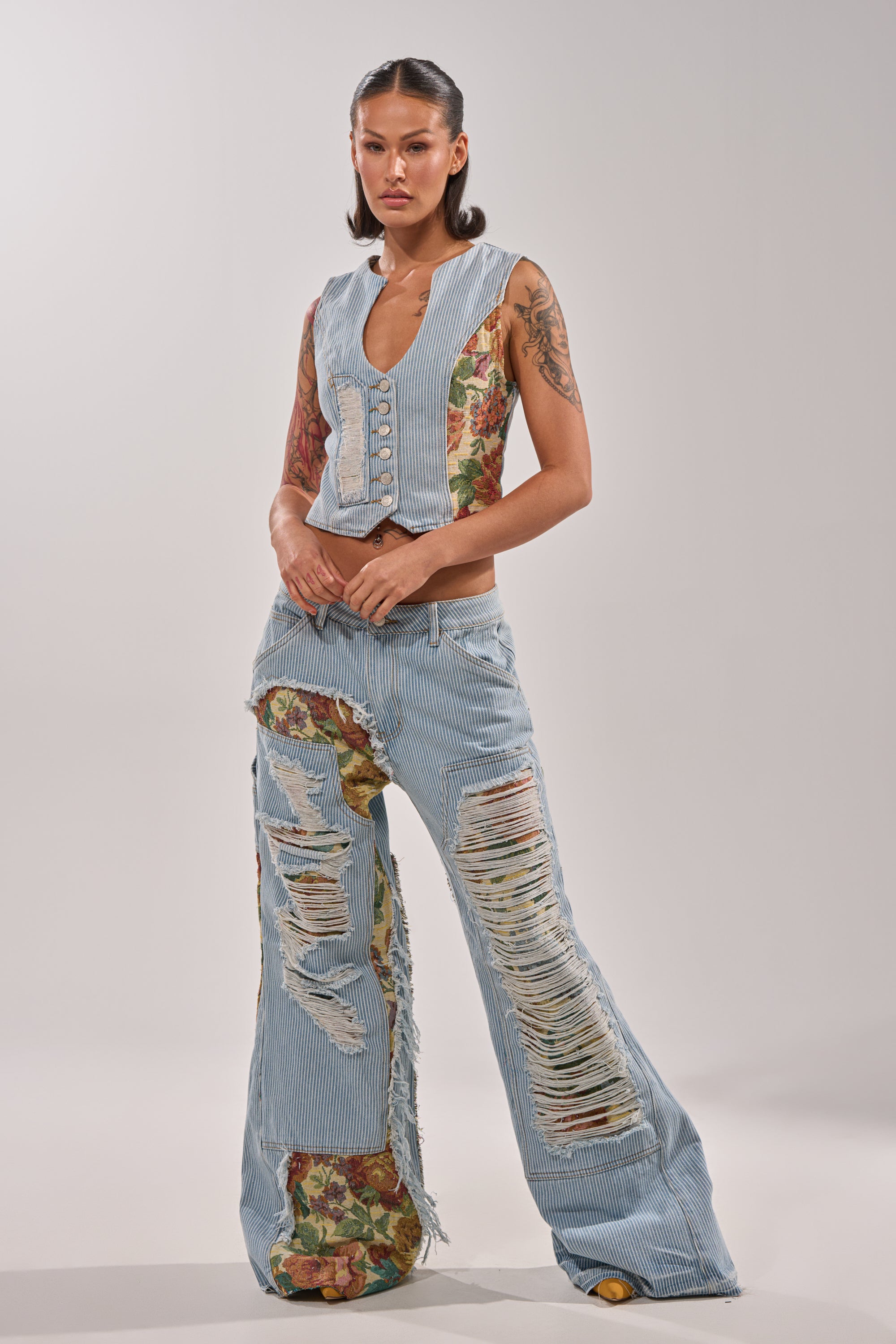 A woman stands confidently in the GUESS WHO STRIPE DENIM WITH BROCADE: a sleeveless, buttoned denim vest and matching distressed wide leg jeans with floral patchwork and striped patterns, set against a plain light background. 5