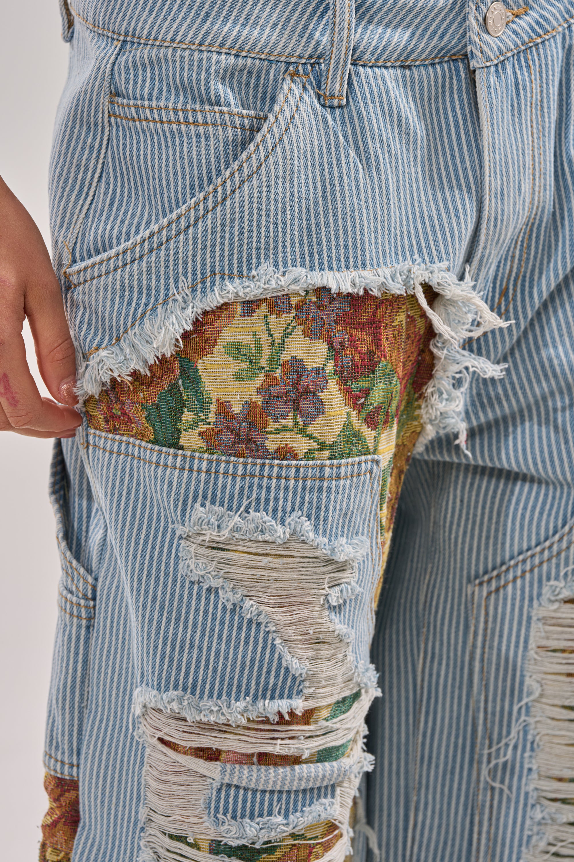 A close-up of the GUESS WHO STRIPE DENIM WITH BROCADE shows wide-leg, blue and white pinstriped jeans with large, frayed rips exposing floral brocade fabric underneath. A left hand rests next to the distressed bottoms. 7