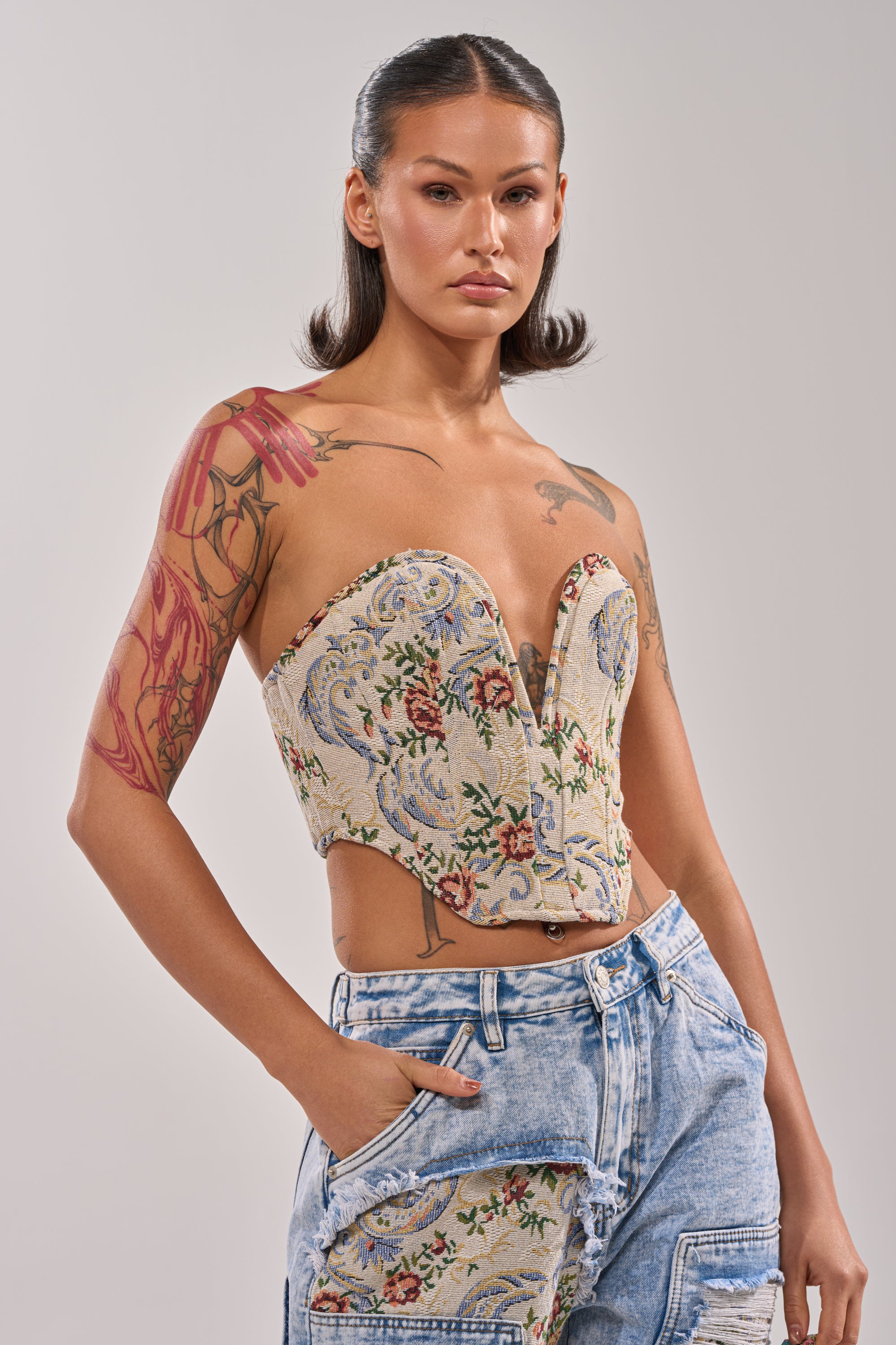A person with long dark hair and floral arm tattoos wears the LANA DISTRESSED JACQUARD CORSET and patchwork denim jeans, hand in pocket, standing against a plain light gray background. 0
