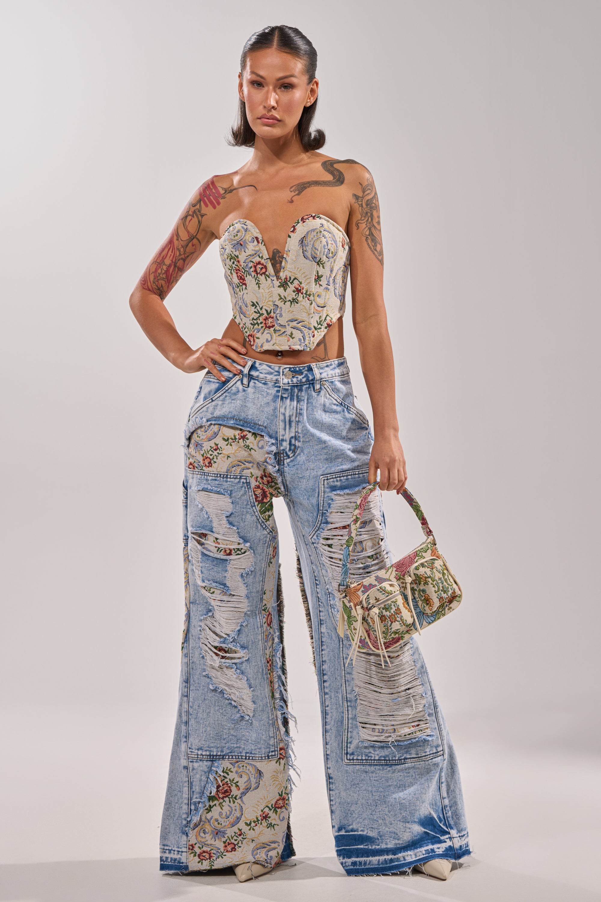 A woman with visible tattoos and sleek hair wears the LANA DISTRESSED JACQUARD CORSET with a strapless sweetheart neckline, paired with distressed wide-leg jeans adorned with floral patches and carries a small matching floral handbag. 4