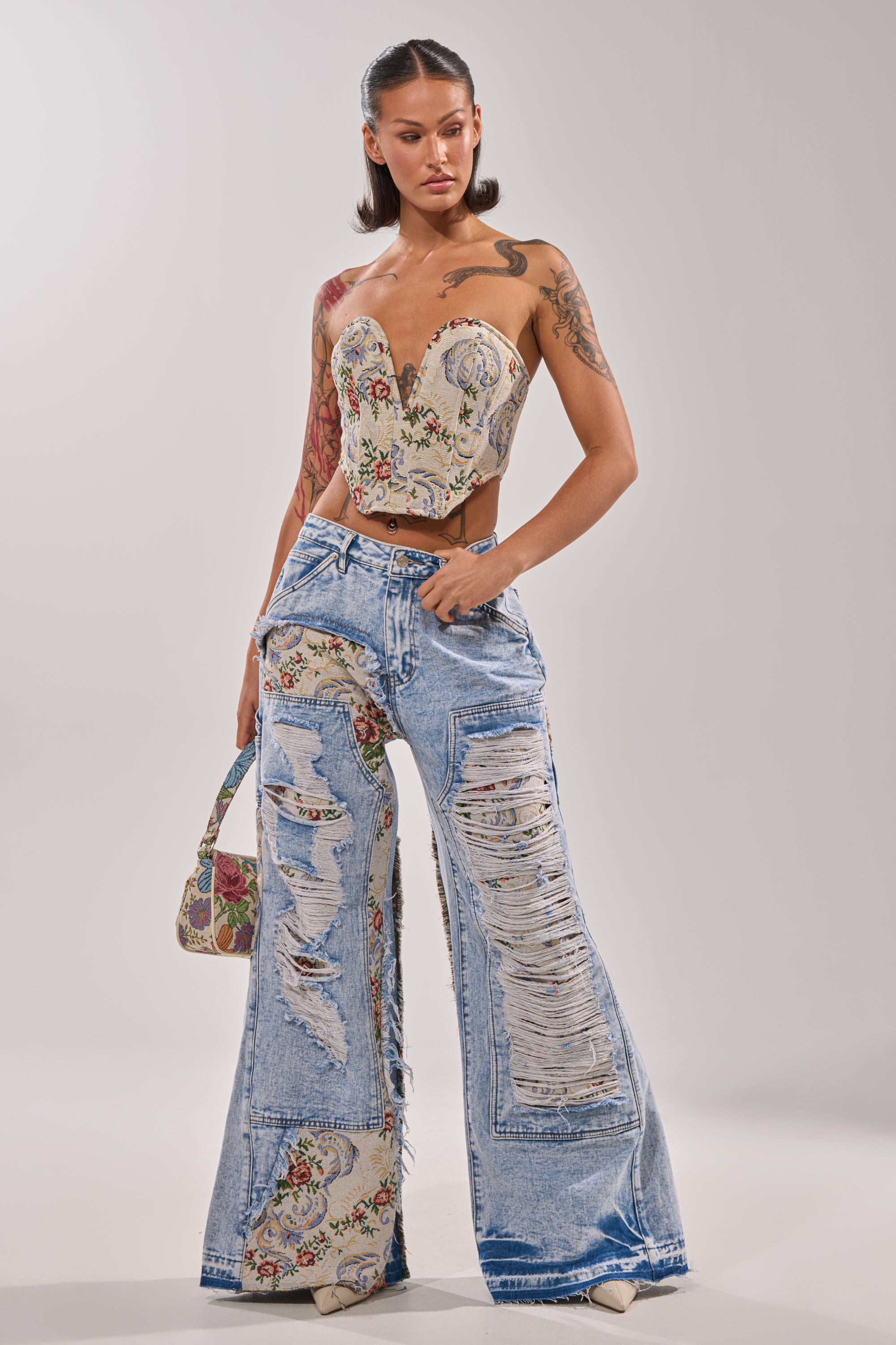 A woman models the LANA DISTRESSED JACQUARD CORSET with a strapless sweetheart neckline, paired with light blue wide-leg ripped jeans featuring floral patches. She accessorizes with a matching floral purse and hoop earrings against a plain background. 5